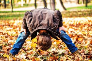 A boy dressed in jeans and a brown jacket bending over and looking at autumn leaves through his legs.