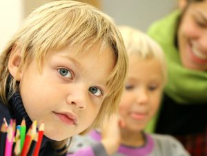 Two young children with a teacher in the background and some colouring pencils in the foreground.