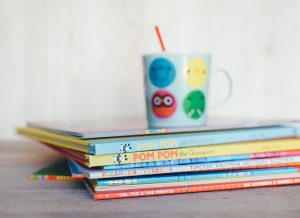 A pile of young children's book with a smiley-face mug on top